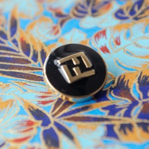 Pode incluir: Um botão preto com um logotipo "E" dourado no centro.