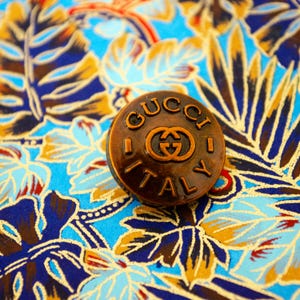 May include: A round, bronze-coloured metal button featuring the embossed text GUCCI at the top and ITALY at the bottom, with the interlocking G logo centred between the words. The button has a dark, weathered finish with raised lettering.