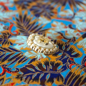 May include: A circular button featuring a central cream-coloured inlay with a gold-toned interlocking CC logo. The edge of the button is framed by a border of small, white, spherical faux pearls set within a scalloped gold-toned metal rim.