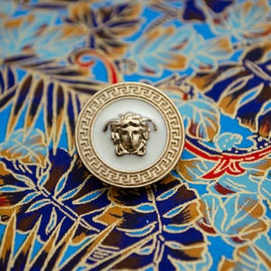 Puede incluir: Un botón dorado con un centro blanco que presenta una cara estilizada. El botón está sobre una tela estampada azul y marrón.