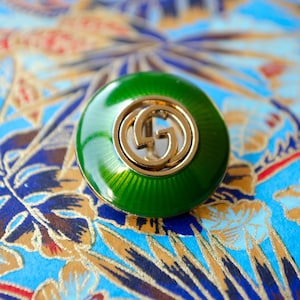 May include: A green enamel button with a gold double-G logo in the center.