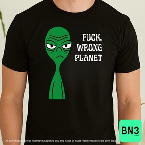 Puede incluir: Camiseta negra con un alienígena verde con expresión de enfado. El texto "FUCK. WRONG PLANET" está impreso en blanco. La camiseta tiene mangas cortas y cuello redondo.