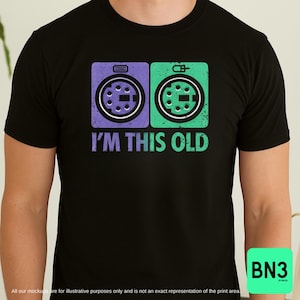 Könnte beinhalten: Schwarzes T-Shirt mit einem Retro-Design mit zwei farbenfrohen Computerport-Symbolen, eines lila und eines grünen, mit dem Text "I'M THIS OLD" in Weiß. Das Shirt hat einen Rundhalsausschnitt und kurze Ärmel.