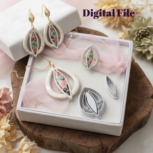 Puede incluir: Un conjunto de joyas con collar, pendientes y anillo con diseño floral. Las piezas blancas en forma de lágrima tienen un borde dorado y un estampado floral en el centro. El conjunto se exhibe en una caja blanca con una tela rosa y el texto "Digital File".