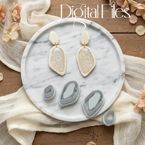 Op de afbeelding: Een paar crèmekleurige, met glitter gevulde oorbellen met een unieke vorm, gepresenteerd op een marmeren dienblad. Er worden ook verschillende grijze polymeerklei-uitsteekvormen in verschillende maten en vormen getoond. De tekst "Digital Files" staat bovenaan.