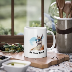 Puede incluir: Taza de cerámica blanca con una llama relajada con gafas de sol, con el texto "No Prob-llama". La llama está representada en estilo acuarela con tonos azules y marrones. La taza está sobre una tabla de cortar de madera.