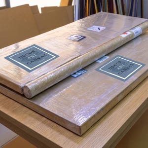 Two packaged flat canvases and one rolled canvas wrapped in clear plastic on a wooden workbench with Decor Art Canvas stickers visible.