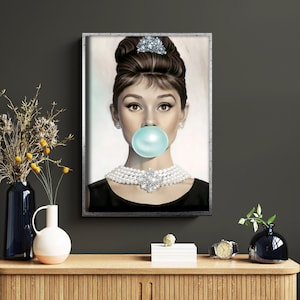 Op de afbeelding: Een ingelijst portret van een persoon met een opgestoken kapsel, een meerrijige parelketting met een centrale kristallen broche en een zwart topje. De persoon blaast een grote, ronde, lichtblauwe kauwgombel voor de mond.