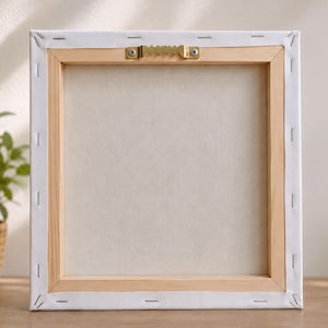 Back of a square stretched canvas with a wooden frame and a brass sawtooth hanger at the top center, showing staples securing the white canvas material.