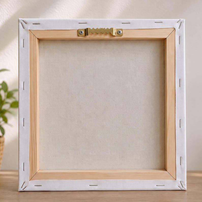 Back of a square stretched canvas with a wooden frame and a brass sawtooth hanger at the top center, showing staples securing the white canvas material.