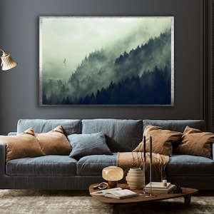 A framed horizontal landscape canvas hangs on a dark gray wall above a blue sofa, depicting a misty forest with tall evergreen trees and a small spaceship flying near a structure.