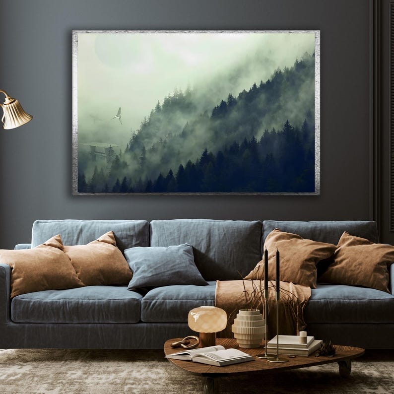 A framed horizontal landscape canvas hangs on a dark gray wall above a blue sofa, depicting a misty forest with tall evergreen trees and a small spaceship flying near a structure.