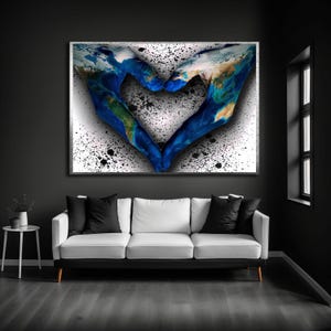 May include: A framed artwork depicts a heart shape formed by two hands, with the Earth's continents painted on them. The background is white with black splatters. The artwork hangs above a white sofa with black pillows.