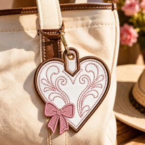 May include: A heart-shaped bag charm featuring white faux leather with pink embroidered scrollwork patterns and a small pink embroidered bow at the bottom. The charm is outlined in brown stitching and attached to a cream canvas tote bag with a gold metal clasp.
