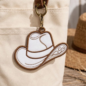 May include: A white embroidered cowboy hat keychain with brown stitched edges and decorative swirl patterns on the brim. The keychain features a gold-toned metal lobster clasp attached to a brown leather strap, hanging from a beige canvas bag.