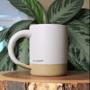 May include: A ceramic mug with a white speckled upper section and a tan speckled base. The mug features a curved handle and the black printed text 03.15.26 followed by a small heart symbol on the lower portion of the white section.