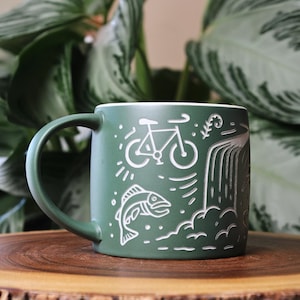 May include: A matte forest green ceramic mug featuring white etched line art illustrations of a bicycle, a jumping fish, and a waterfall. The mug has a smooth, cylindrical body with a curved handle on the left side and a white interior rim.