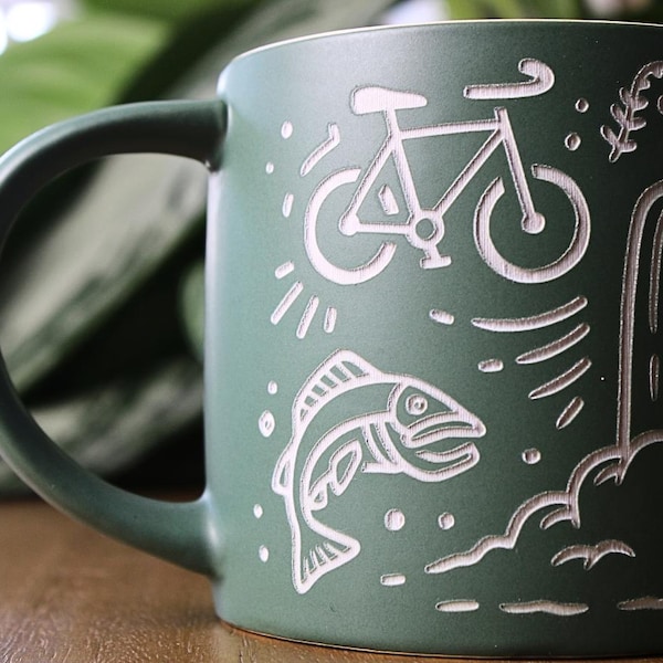 Könnte beinhalten: Ein matter, waldgrüner Keramikbecher mit weißem Gravurdesign. Die Illustration zeigt ein Fahrrad, einen springenden Fisch und einen Wasserfall mit stilisierten Wassertropfen sowie ein Farnblatt-Detail an der Seite.