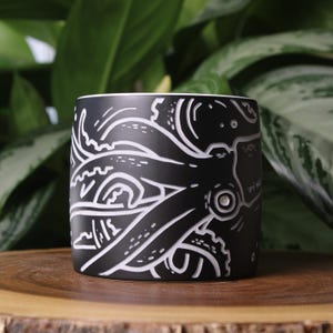 May include: Black ceramic planter with a white octopus design. The planter features a cylindrical shape with a white rim and a detailed illustration of an octopus wrapping around the exterior. The planter is set on a wooden surface.