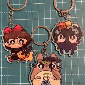 May include: Three acrylic keychains featuring anime-style characters. One keychain depicts a girl with a black cat, another a dark creature with flower accents, and the third a gray creature holding an umbrella with a smaller creature.