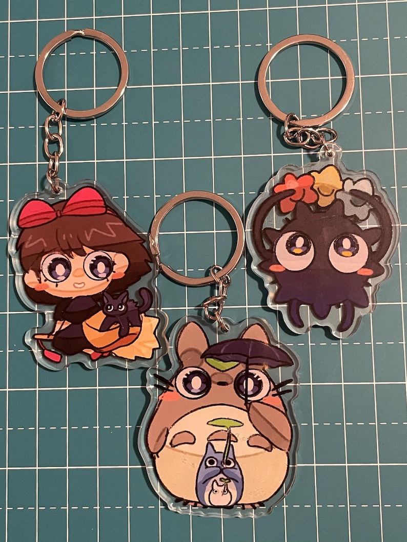 May include: Three acrylic keychains featuring anime-style characters. One keychain depicts a girl with a black cat, another a dark creature with flower accents, and the third a gray creature holding an umbrella with a smaller creature.