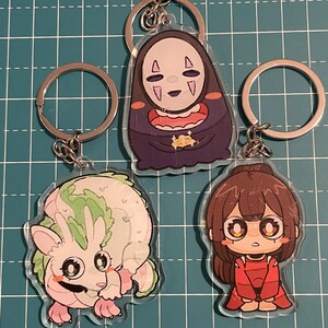 May include: Three anime-style keychains with silver key rings. One keychain features a character in a dark purple robe with a white mask. Another depicts a pink and green creature. The third shows a girl in a red outfit.
