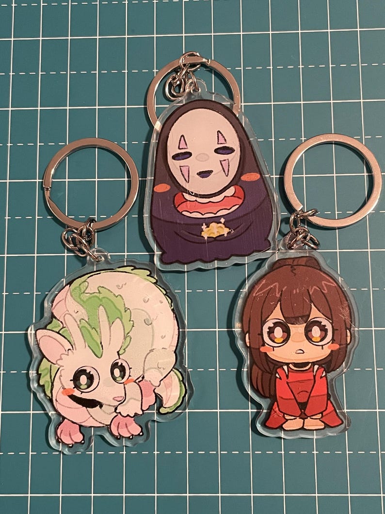May include: Three anime-style keychains with silver key rings. One keychain features a character in a dark purple robe with a white mask. Another depicts a pink and green creature. The third shows a girl in a red outfit.