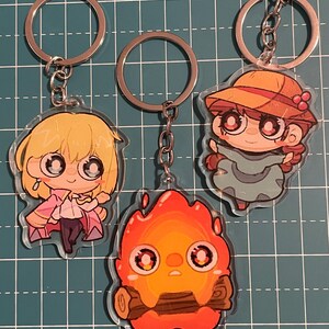 May include: Three anime-style keychains featuring characters with large eyes. One keychain depicts a blonde character in a pink outfit, another shows a fiery creature, and the third features a character in a hat. Each keychain has a silver keyring.