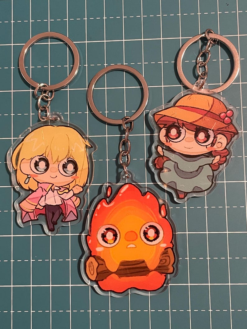May include: Three anime-style keychains featuring characters with large eyes. One keychain depicts a blonde character in a pink outfit, another shows a fiery creature, and the third features a character in a hat. Each keychain has a silver keyring.