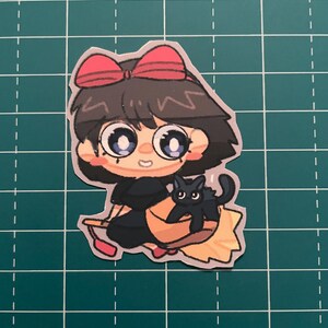以下が含まれることがあります： 赤いリボン、黒いドレス、ほうきに乗った黒猫が描かれた漫画の魔女のステッカー。魔女は大きな目をし、微笑んでいます。ほうきは黄色とオレンジ色です。