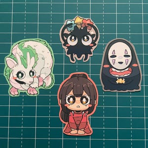 以下が含まれることがあります： アニメスタイルのカラフルなステッカー5枚のコレクション。緑色のアクセントが付いた白い生き物、花の冠をつけた黒い生き物、赤い衣装のキャラクターなど、さまざまなデザインのキャラクターが描かれています。