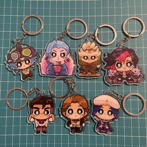 May include: Collection of acrylic keychains featuring cartoon characters. Each keychain has a silver-colored ring and chain. The characters have various hair colors and outfits, with detailed facial features and expressions. The keychains are arranged on a green grid surface.
