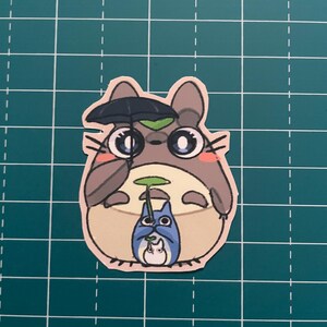 以下が含まれることがあります： 傘と葉っぱを持った、漫画のトトロのキャラクターのステッカー。トトロはグレーとタンで、大きな目と笑顔の表情をしています。小さな青いトトロと白いトトロも登場します。ステッカーはピンク色で縁取られています。