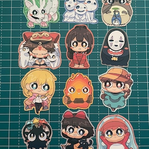 以下が含まれることがあります： 様々なキャラクターが描かれた、カラフルなアニメスタイルのステッカーのコレクション。ステッカーには、ドラゴン、白い精霊のグループ、トトロなど、様々なデザインが鮮やかな色で表現されています。ステッカーは緑色のグリッド上に配置されています。