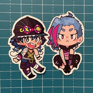 Puede incluir: Dos pegatinas de personajes de dibujos animados, una con el pelo azul y una raya rosa que lleva un atuendo negro y rosa, y la otra con el pelo azul y un sombrero negro que lleva un atuendo negro y amarillo.