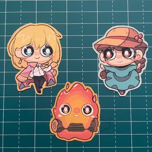 以下が含まれることがあります： 人気アニメ映画のキャラクターをフィーチャーした3つの漫画ステッカー。ステッカーは、ピンクの衣装を着たブロンドのキャラクター、青いドレスと帽子を着たキャラクター、丸太を持った燃えるような生き物を描いています。