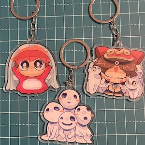 May include: Three anime-style keychains with silver key rings. One keychain features a red and white character in a bubble. Another shows a character with a hat and two small white creatures. The third keychain has a group of white, smiling characters.