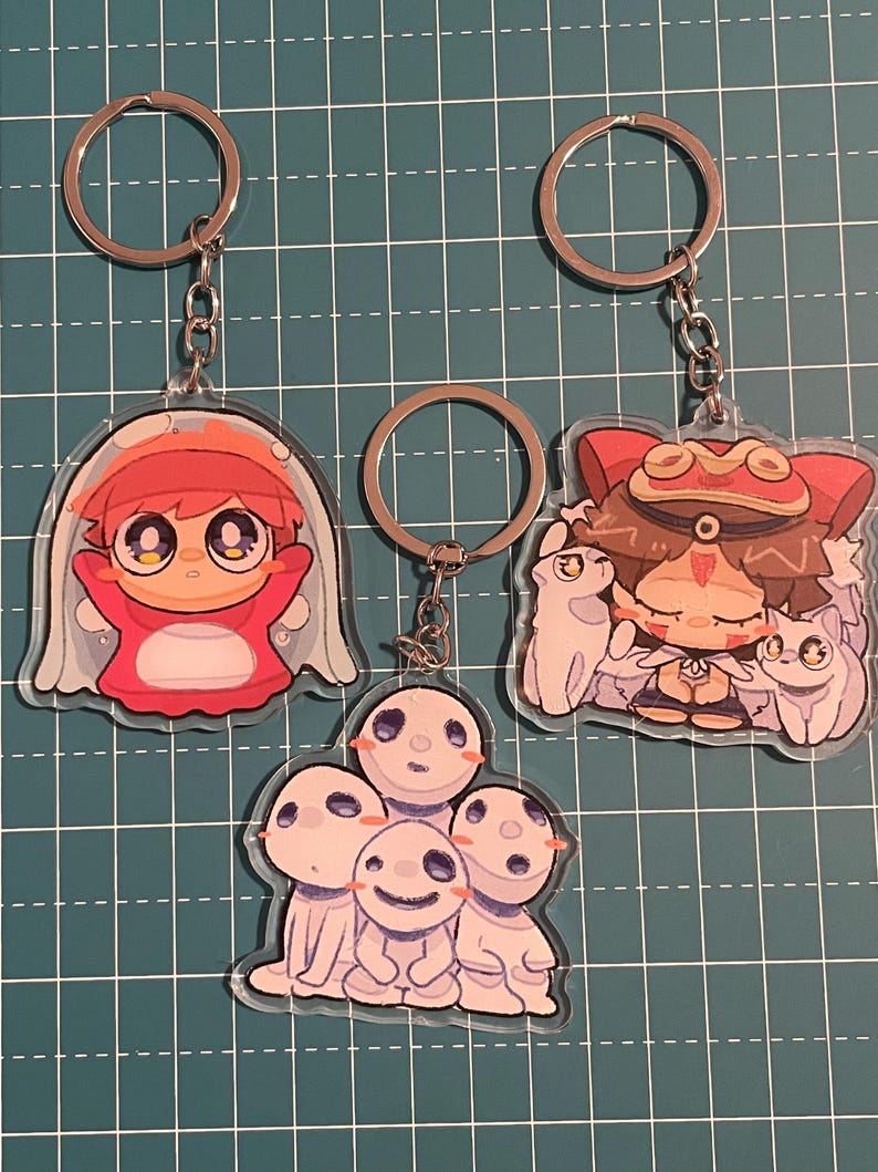 May include: Three anime-style keychains with silver key rings. One keychain features a red and white character in a bubble. Another shows a character with a hat and two small white creatures. The third keychain has a group of white, smiling characters.