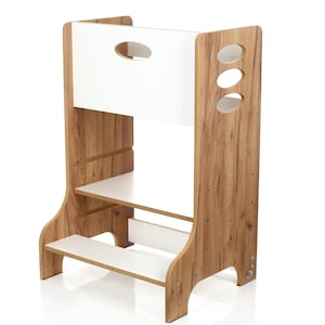 Op de afbeelding: Een houten opstapje met een witte bovenkant en planken. De zijkanten hebben een houtnerf afwerking. Het bovenste gedeelte heeft een ovale uitsparing voor een handvat. Het zijpaneel heeft drie ovale uitsparingen. De kruk heeft twee planken en een voetensteun.