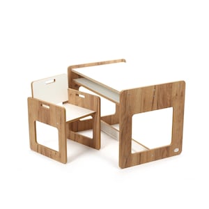 Op de afbeelding: Een kleine houten bureau- en stoelset voor kinderen. Het bureau heeft een wit tafelblad en een onderste plank. De stoel heeft een witte zitting en rugleuning. Het hout heeft een lichtbruine houtnerf.