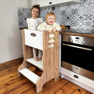 Può includere: Uno sgabello da cucina in legno con finitura bianca e venature del legno. Lo sgabello ha tre livelli ed è progettato per aiutare i bambini a raggiungere i piani di lavoro. Due bambini sono in piedi sullo sgabello.