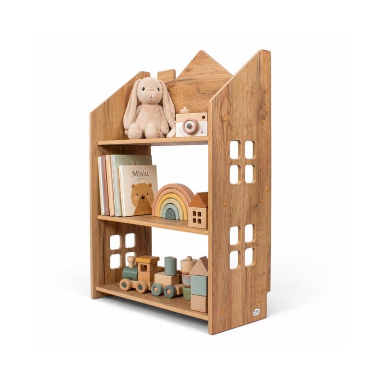 May include: A light brown wooden house-shaped bookshelf with three shelves. The top shelf holds a plush rabbit and a toy camera. The middle shelf contains books, a wooden rainbow, and a house block. The bottom shelf features a wooden toy train.