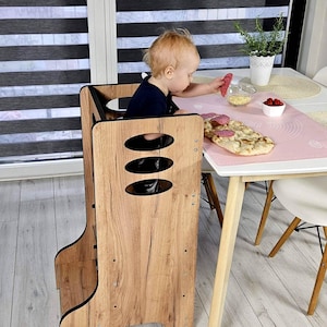 Pode incluir: Uma torre de aprendizagem de madeira para crianças pequenas, com acabamento castanho claro. A torre tem três recortes ovais e uma base curva. Uma criança está sentada na torre, interagindo com comida numa mesa próxima.