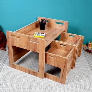 Puede incluir: Un conjunto de mesa y dos sillas de madera para niños. La mesa tiene una bandeja superior y las sillas tienen compartimentos de almacenamiento. El conjunto está hecho de madera marrón claro y está sobre una alfombra de patrón blanco y gris.