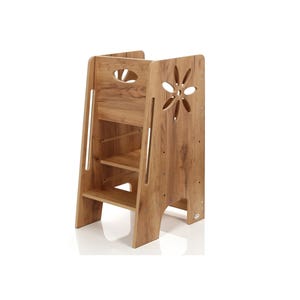 Op de afbeelding: Een houten opstapje met een natuurlijke houtnerf afwerking. De kruk heeft twee treden en een uitgesneden bloemmotief op het zijpaneel. Het bovenpaneel heeft een handgreepvormige uitsparing. De kruk is ontworpen voor kinderen.