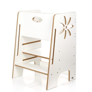 Può includere: Uno sgabello in legno bianco con un ritaglio a forma di fiore sul lato. Lo sgabello ha tre gradini e un ritaglio per la maniglia nella parte superiore. I bordi dello sgabello sono di colore legno naturale.