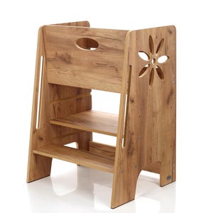 Può includere: Uno sgabello in legno con finitura a venatura del legno marrone chiaro. Lo sgabello ha due gradini e una piattaforma superiore con un ritaglio ovale. Un ritaglio a forma di fiore decora il pannello laterale. Lo sgabello è per bambini.