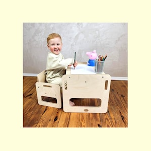Op de afbeelding: Een lichtgekleurde houten kinderstoel en -tafelset. De tafel heeft een rechthoekig blad met een uitsparing voor de benen. Een kind zit aan de tafel en houdt een potlood vast. Een roze varkensbeeldje en een beker met kleurpotloden staan op de tafel.