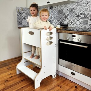 Può includere: Uno sgabello da cucina in legno bianco con due bambini in piedi sopra. Lo sgabello ha un design rettangolare con bordi arrotondati e ritagli ovali. I bambini sorridono e guardano la fotocamera. Lo sfondo include una cucina con un paraschizzi a motivi.