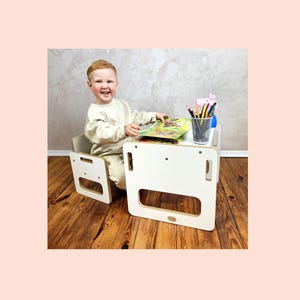 Op de afbeelding: Een witte houten tafel en stoel set voor kinderen. De tafel heeft een rechthoekig blad met aan elke kant een uitgesneden handvat. De stoel heeft een vergelijkbaar ontwerp. Het kind zit aan de tafel en leest een boek. Een bakje met potloden en stiften staat op de tafel.
